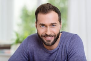 Head and shoulders portrait of a smiling bearded man looking at the camera with a friendly smile, indoors at home